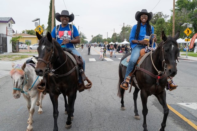 A man and woman of color, wearing cowboy hats, riding horses down a streets closed to car traffic. The man walk a small pony next to his horse.
